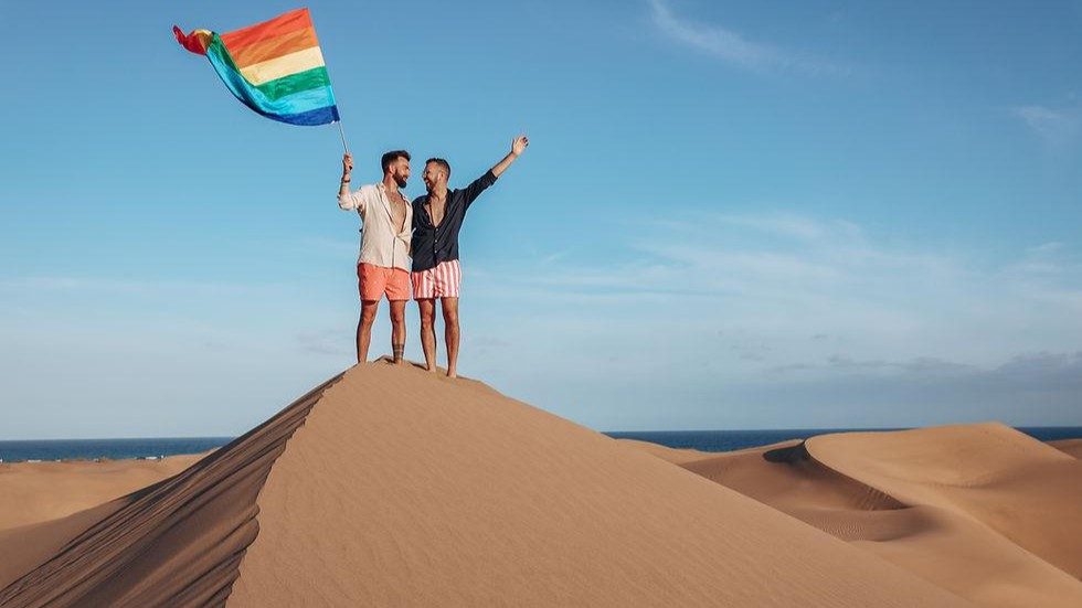 Es sind zwei Männer die eine Regenbogenfahne hochhalten und zeigen  einen Sandberg in den Dünen von Maspalomas auf Gran Canaria